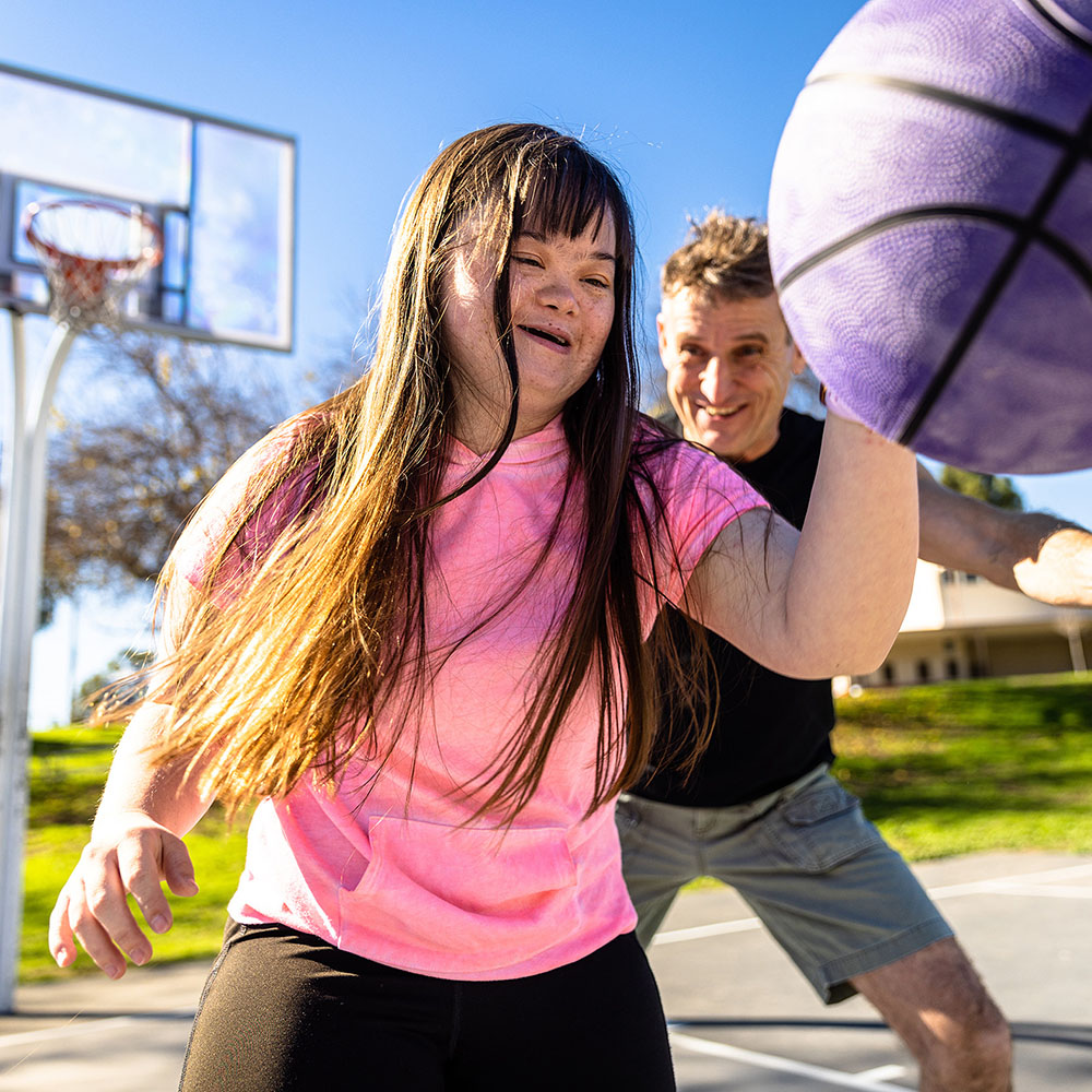 A young girl wearing a pink shirt plays basketball with her disability support worker