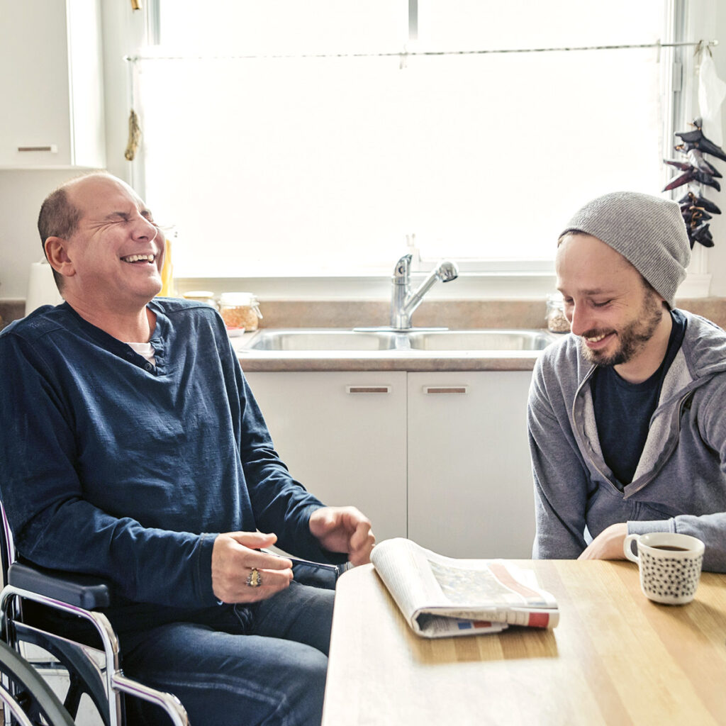 Two men sit in a kitchen enjoying coffee together. One of the men is in a wheelchair. Both are laughing.